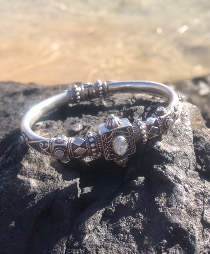 K&ouml;nnte beinhalten: Ein silberfarbenes Armband mit aufwendigen Details, das ein zentrales quadratisches Element mit einem klaren Stein aufweist. Das Armband liegt auf einem dunklen, strukturierten Felsen mit verschwommenem Wasserhintergrund, was sein kompliziertes Design und seine Handwerkskunst zeigt.