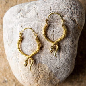 Könnte beinhalten: Ein Paar goldfarbene Creolen mit Schlangenmuster. Die Ohrringe sind strukturiert und haben unten einen Schlangenkopf. Die Ohrringe werden auf einem hellgrauen Stein präsentiert. Die Ohrringe sind ein Schmuckstück.
