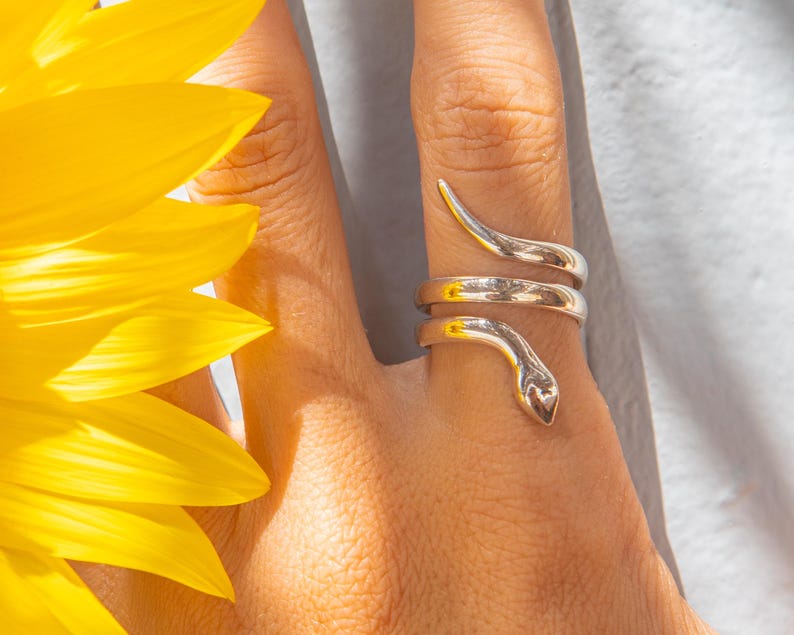 Puede incluir: Un anillo plateado con forma de serpiente que se enrolla alrededor de un dedo. El dise&ntilde;o del anillo presenta una cabeza y una cola de serpiente, con el cuerpo de la serpiente enrollado alrededor del dedo. El anillo se lleva en una mano, con un girasol amarillo en el fondo.