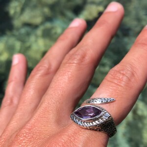 May include: A silver snake ring with a purple amethyst gemstone. The ring is on a person's finger.