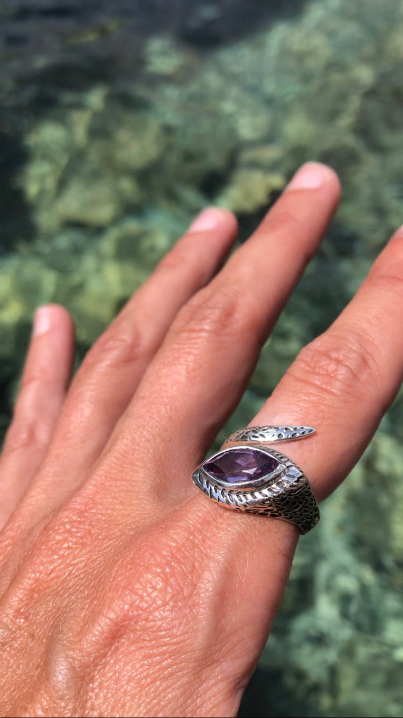 May include: A silver snake ring with a purple amethyst gemstone. The ring is on a person's finger.