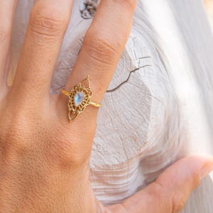 Peut inclure: Une bague dorée avec un motif filigrané orné. La bague présente une pierre précieuse irisée en forme de poire au centre. La bague est portée sur un doigt, mettant en valeur ses détails délicats et son savoir-faire. C'est un bijou.