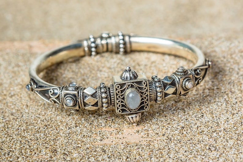 K&ouml;nnte beinhalten: Silberfarbenes Armreif-Armband mit aufwendigen Details. Das Armband hat ein quadratisches Mittelelement mit einem ovalen, hellfarbigen Stein, umgeben von komplizierten Mustern und kleineren, runden Steinen. Es liegt auf einer sandigen Oberfl&auml;che.