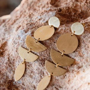 May include: Gold-toned earrings with a geometric design. Each earring features a small circle at the top, connected to an oval, a half-circle, and a teardrop shape. The earrings are displayed on a textured, brown surface.