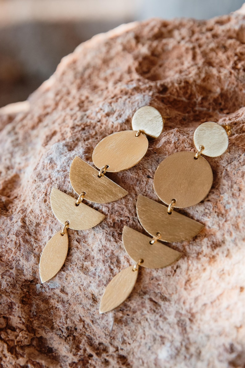 May include: Gold-toned earrings with a geometric design. Each earring features a small circle at the top, connected to an oval, a half-circle, and a teardrop shape. The earrings are displayed on a textured, brown surface.