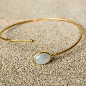 Puede incluir: Un brazalete de oro con una piedra ovalada blanca. El brazalete está sobre una superficie arenosa.
