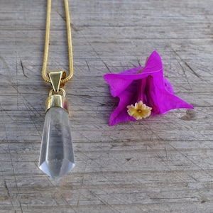 May include: A gold-colored chain necklace with a faceted, clear crystal pendant. The pendant is a light gray color. A bright pink flower with a yellow center is next to the necklace on a wooden surface.