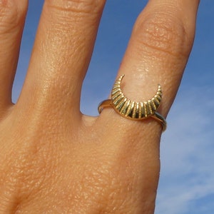 May include: A gold crescent moon ring with a textured design. The ring is worn on a finger against a blue sky background.