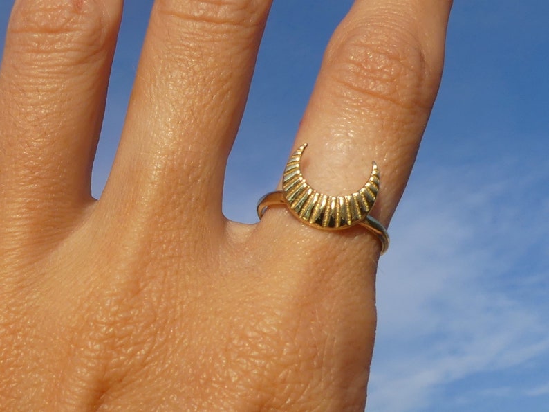 May include: A gold crescent moon ring with a textured design. The ring is worn on a finger against a blue sky background.