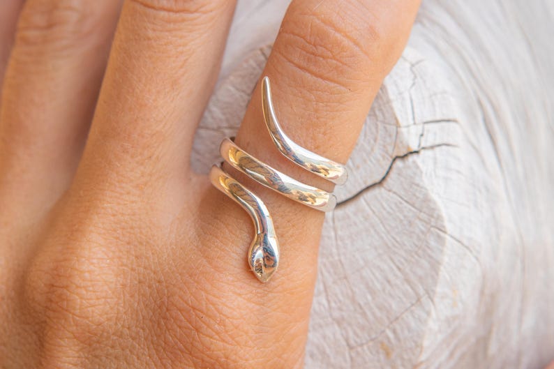 Puede incluir: Un anillo de serpiente plateado envuelve un dedo. El anillo presenta un dise&ntilde;o de cabeza y cola de serpiente, con el cuerpo de la serpiente enroll&aacute;ndose alrededor del dedo en espiral. El anillo es de metal brillante.