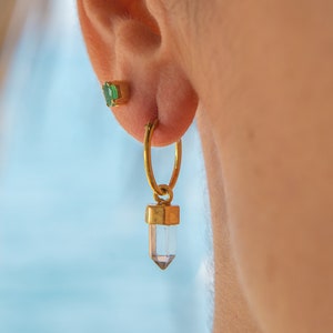 May include: Gold hoop earrings with a clear crystal pendant. The crystal is faceted and has a gold setting. The earrings are worn in a person's ear.