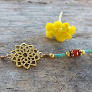 May include: A delicate gold-colored necklace featuring a central openwork floral pendant. The necklace incorporates small turquoise beads, gold-colored beads, and a red bead. A yellow flower rests in the background.