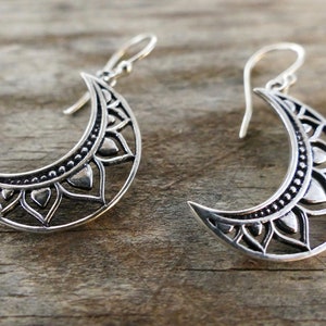May include: Silver crescent moon earrings with intricate floral and dot patterns. The earrings have a hook closure and are displayed on a wooden surface. These earrings are a unique accessory.