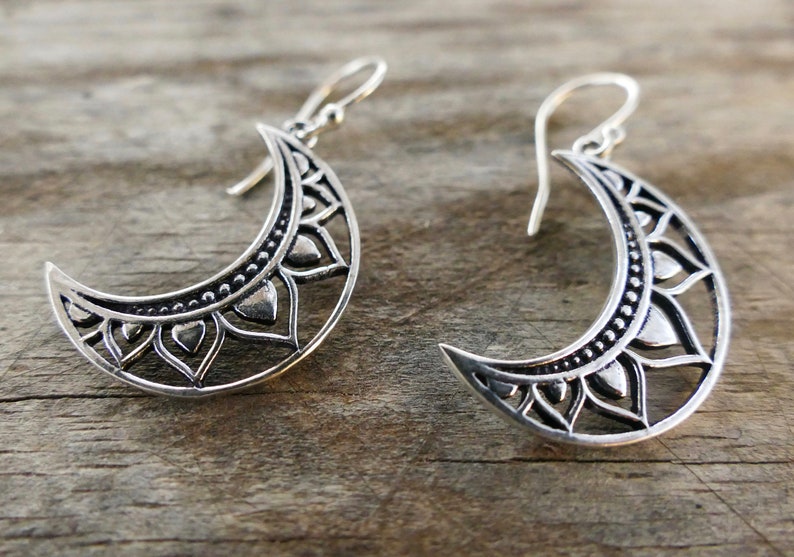 May include: Silver crescent moon earrings with intricate floral and dot patterns. The earrings have a hook closure and are displayed on a wooden surface. These earrings are a unique accessory.
