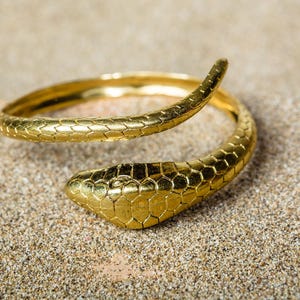 Puede incluir: Un anillo de serpiente dorado con un patrón texturizado en forma de escamas. El anillo está enrollado, con la cabeza de la serpiente en un extremo y la cola en el otro. La joya está sobre un fondo arenoso.