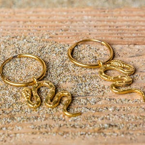 Op de afbeelding: Een paar gouden creolen met een slangenbedel aan elke oorring. De slangenbedels zijn gedetailleerd met schubben en hebben een gekrulde staart.
