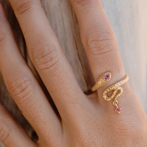 Op de afbeelding: Een goudkleurige slangenring met een getextureerd oppervlak. De kop en staart van de slang wikkelen zich om de vinger, met een kleine roze edelsteen op de kop en een hangende roze edelsteen op de staart.