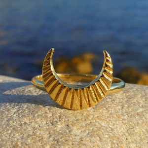 May include: A gold crescent moon ring with a textured design. The ring is on a light brown rock.