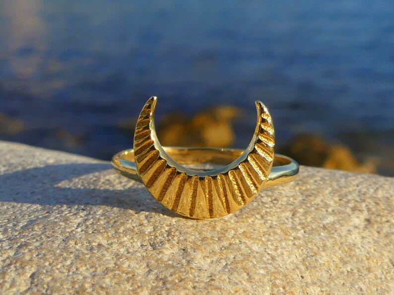 May include: A gold crescent moon ring with a textured design. The ring is on a light brown rock.