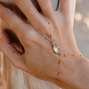 Könnte beinhalten: Eine goldfarbene Handkette bestehend aus einer feinen Gliederkette mit kleinen, gleichmäßig verteilten Kügelchen. Das Schmuckstück verbindet einen Ring mit einem Armband und ist mittig mit einem tropfenförmigen, durchscheinenden Stein besetzt.