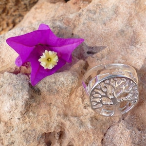 Puede incluir: Un anillo de plata con un diseño de árbol de la vida. El anillo está sobre una roca con una flor morada.