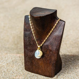 Può includere: Una collana color oro con un ciondolo con una pietra azzurra e un ciondolo a forma di mezzaluna dorata. La collana è esposta su un espositore di gioielli in legno marrone scuro, su uno sfondo sabbioso.