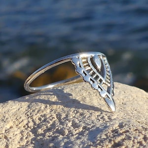 Könnte beinhalten: Ein silberner Ring mit einem filigranen, komplizierten Design in Form eines Dreiecks. Der Ring ist vor dem Hintergrund einer felsigen Oberfläche und verschwommenem blauen Wassers platziert.