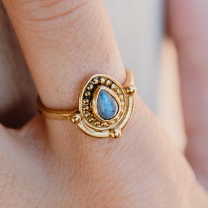 Puede incluir: Un anillo dorado con una piedra preciosa azul en forma de lágrima. El anillo tiene un diseño detallado con cuentas alrededor de la piedra y en la banda. El anillo es una joya.