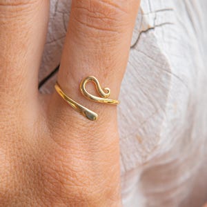 Può includere: Un anello color oro con un design unico e aperto. L'anello si avvolge attorno al dito con una texture martellata su un'estremità e un ricciolo decorativo sull'altra. L'anello è mostrato su una mano.