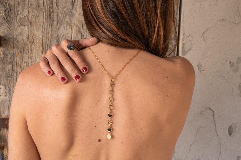 May include: A gold chain necklace with a series of small, round, gold charms hanging from it. The necklace is worn on the back of the neck, with the charms hanging down the back.