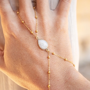 Puede incluir: Una cadena de mano dorada con un colgante de piedra preciosa blanca en forma de pera. La cadena presenta pequeñas cuentas redondas y eslabones delicados, creando un diseño elegante y minimalista. La joya se muestra en una mano.