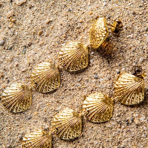 Puede incluir: Un par de pendientes dorados con forma de concha marina sobre arena. Los pendientes presentan una serie de colgantes conectados en forma de abanico, cada uno con vetas detalladas. Los pendientes están diseñados para colgar.