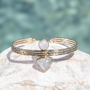 May include: Gold-coloured cuff bracelet with a textured surface. The bracelet features a round and a diamond-shaped moonstone. The jewellery is displayed on a white rock with a blurred turquoise background.