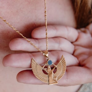 May include: Gold-toned pendant necklace featuring an Egyptian goddess design with outstretched wings and a blue gemstone. The pendant hangs from a delicate chain. The necklace is held in a person's hand.