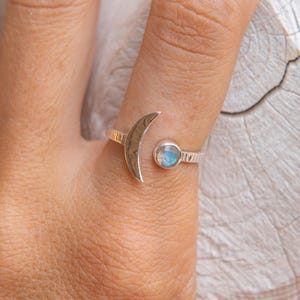 Puede incluir: Un anillo de plata con un diseño de media luna y una pequeña piedra redonda de color azul claro. El anillo se lleva en un dedo, mostrando la joya de temática celestial. La banda del anillo tiene un patrón texturizado.
