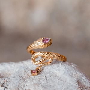May include: A gold snake ring with a textured pattern. The snake's head and tail have pink gemstones. The ring is shown on a light-coloured stone surface.