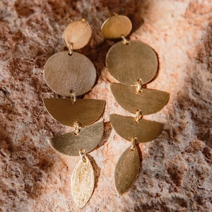 Könnte beinhalten: Goldfarbene Hängeohrringe mit einem geschichteten Design. Jeder Ohrring hat oben einen kleinen Kreis, gefolgt von einem größeren Kreis, zwei Halbkreisformen und einem blattförmigen Anhänger. Die Ohrringe werden auf einer strukturierten, rotbraunen Oberfläche präsentiert.