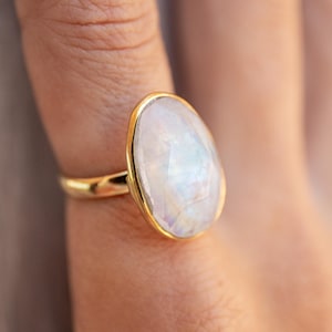 Puede incluir: Un anillo de oro con una gran piedra de luna ovalada. La piedra es iridiscente y tiene un color blanco lechoso.