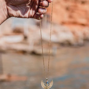 May include: A gold-colored necklace with a pendant featuring a winged figure. The pendant has blue and red accents. The necklace is held up against a blurred background of earth tones.