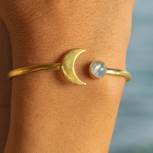 Puede incluir: Brazalete de puño dorado con una luna creciente y una piedra preciosa redonda e iridiscente. El brazalete se lleva en un brazo, mostrando su diseño celestial. El fondo es de color azul suave.