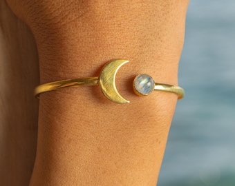 May include: Gold-toned cuff bracelet featuring a crescent moon and a round, iridescent gemstone. The bracelet is worn on a person's arm, showcasing its celestial design. The background is a soft blue.