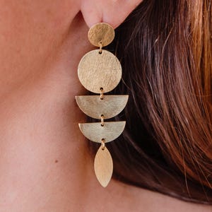 May include: Gold-toned geometric earrings with a brushed finish. The earrings feature a circular stud, a large circle, two half-circle shapes, and a teardrop-shaped dangle. The earrings are connected by small loops.