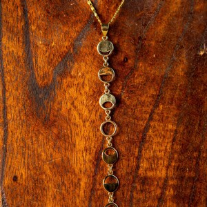 May include: A gold necklace with a pendant made of multiple connected circles. The circles are arranged in a vertical line and are of varying sizes. The necklace is displayed on a wooden surface.