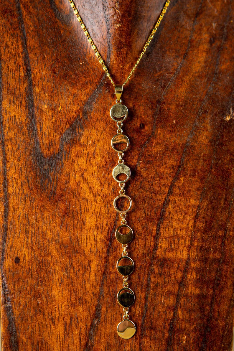 May include: A gold necklace with a pendant made of multiple connected circles. The circles are arranged in a vertical line and are of varying sizes. The necklace is displayed on a wooden surface.