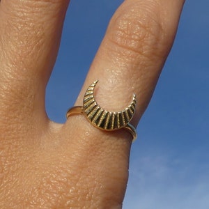 May include: A gold crescent moon ring with a textured surface. The ring is worn on a finger.