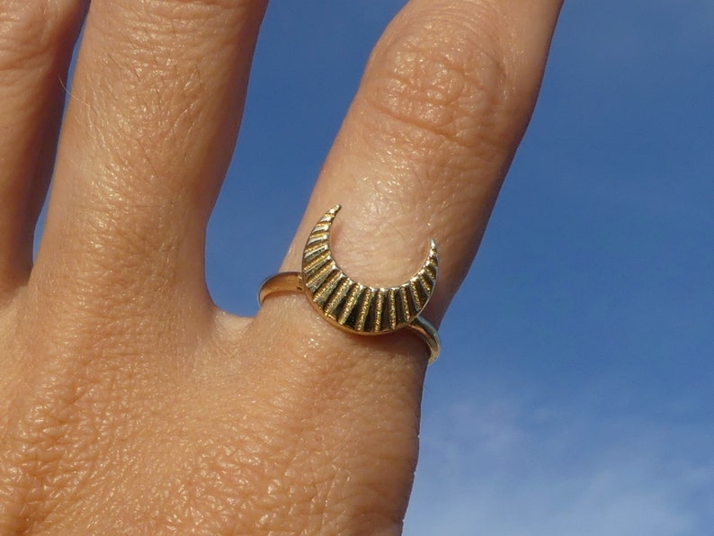 May include: A gold crescent moon ring with a textured surface. The ring is worn on a finger.