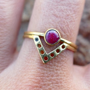 May include: Gold ring with a red gemstone and a geometric design. The ring is made of two bands, one with a V-shaped design and the other with a circle of holes.