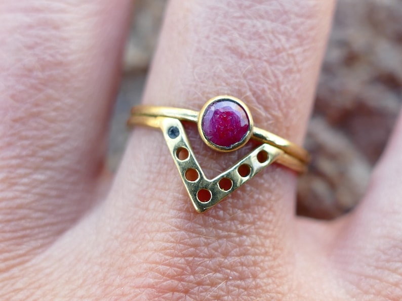 May include: Gold ring with a red gemstone and a geometric design. The ring is made of two bands, one with a V-shaped design and the other with a circle of holes.