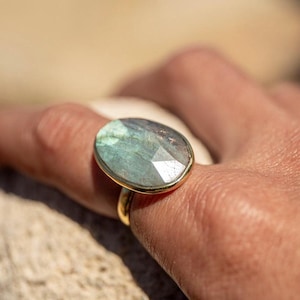 May include: A gold ring featuring a large, oval-shaped, iridescent green gemstone. The ring is worn on a finger.