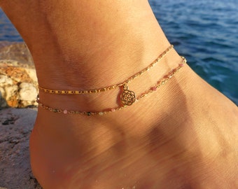 May include: Gold anklet with a delicate chain and a small charm in the shape of a flower. The anklet is layered with a second chain that has small pink and green beads.